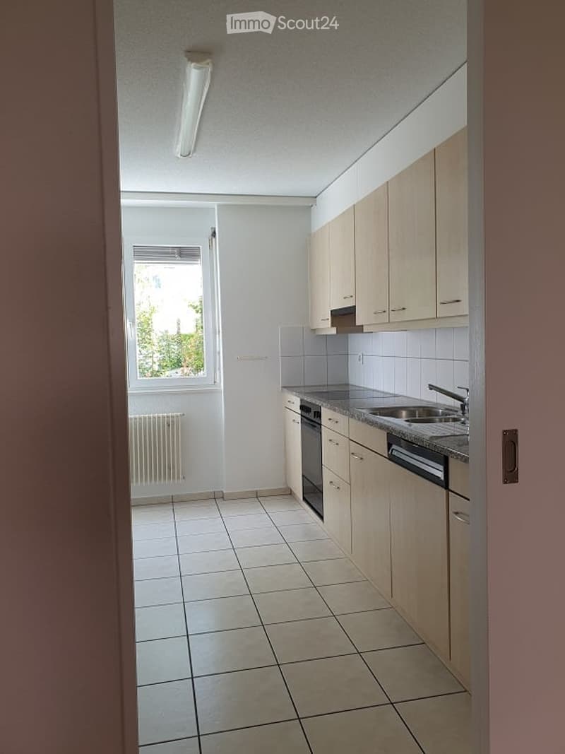 Modern kitchen with light wooden cabinets, tiled floor, and a window providing natural light. Includes a sink, faucet, oven, and radiator.