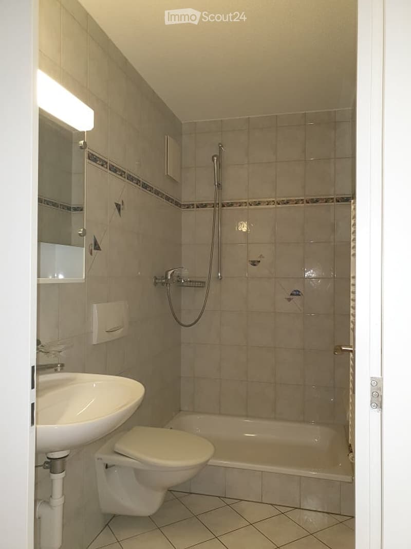 Bathroom with beige tiled walls and floor, featuring a sink, toilet, bathtub, and shower. A mirror is mounted on the wall above the sink, accompanied by a light fixture.