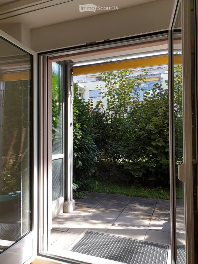 Open sliding glass door leading to a small balcony or terrace area with tiled flooring and a metal drain cover. Greenery and neighboring buildings are visible in the background.