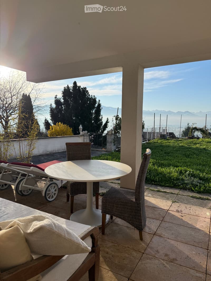 Balcony with a round table, two chairs, and a sun lounger, overlooking a scenic view of trees and mountains.