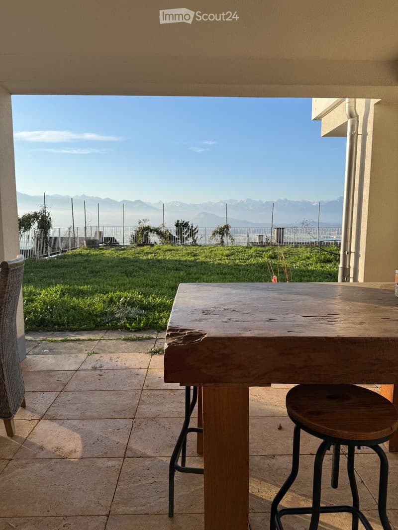Balcony with wooden table, stool, and chair, overlooking a grassy area and distant mountains.