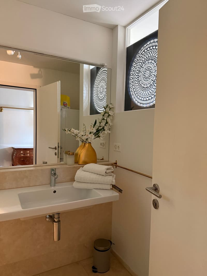 Modern bathroom with a white sink, chrome faucet, large mirror, neatly folded towels, decorative vase with white flowers, and a trash can.
