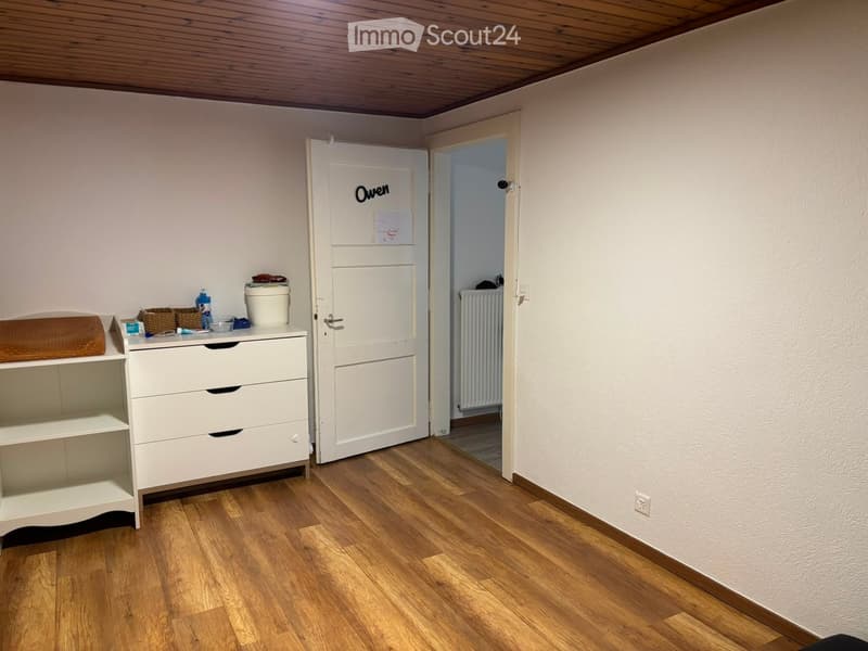A small, furnished room with wooden flooring, a white cabinet with drawers, a shelf, and a door labeled "Owen" leading to another room.