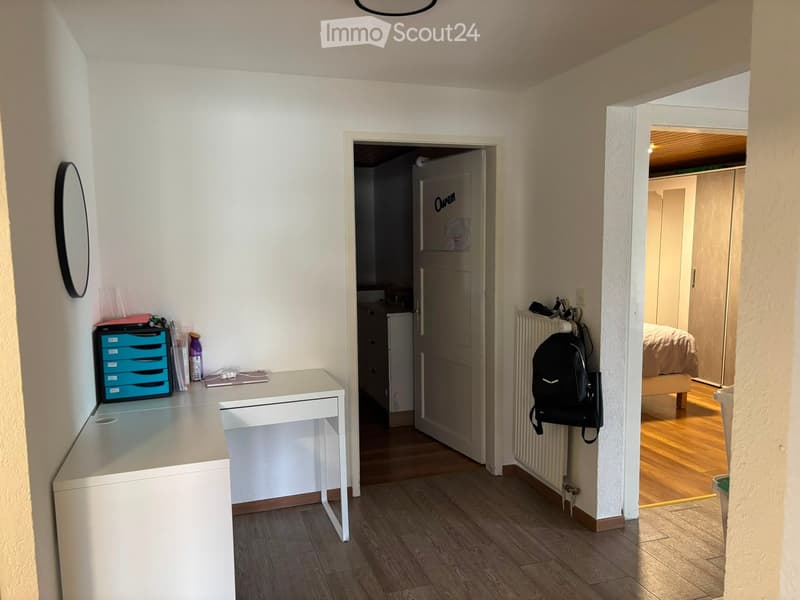 A narrow hallway with light wooden flooring, white walls, and several doors leading to different rooms. A small desk with a blue drawer unit and a mirror is visible on the left, while a backpack hangs on the right wall. A bedroom with a bed and wardrobe i