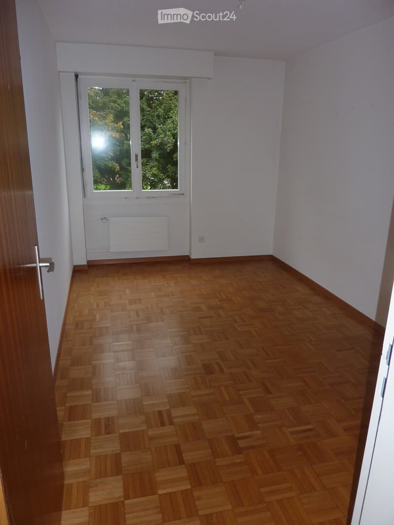Empty room with wooden parquet flooring, white walls, and a large window with white frames and blinds. A radiator is visible below the window, and a door with a silver handle is on the left side.