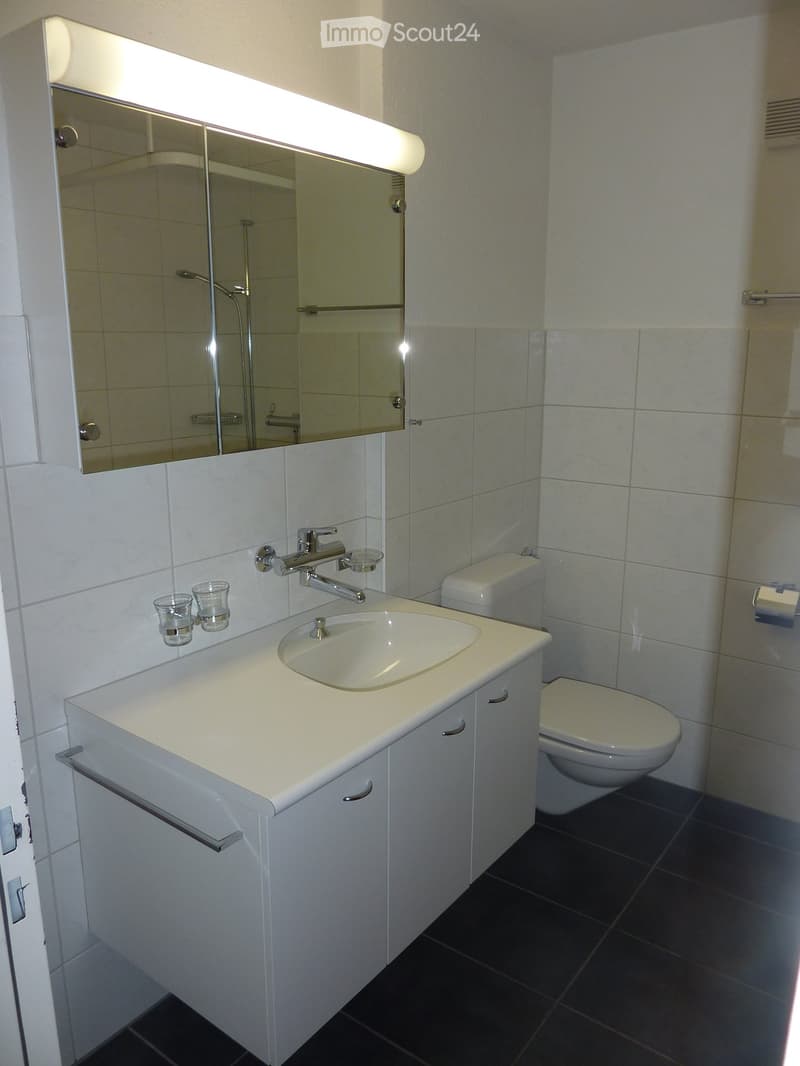 Modern bathroom with white tiles, a sink with a modern faucet, a mirror with built-in lighting, a toilet, and a small cabinet under the sink.