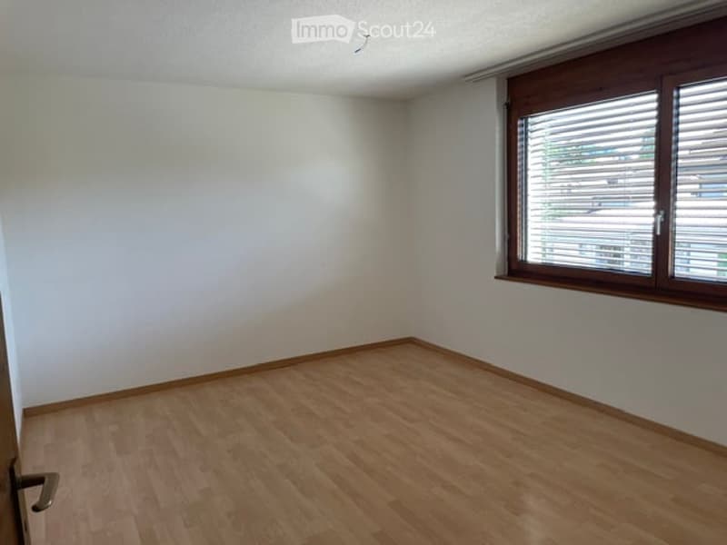 Empty room with light wooden flooring and white walls, featuring a large window with wooden frame and blinds.