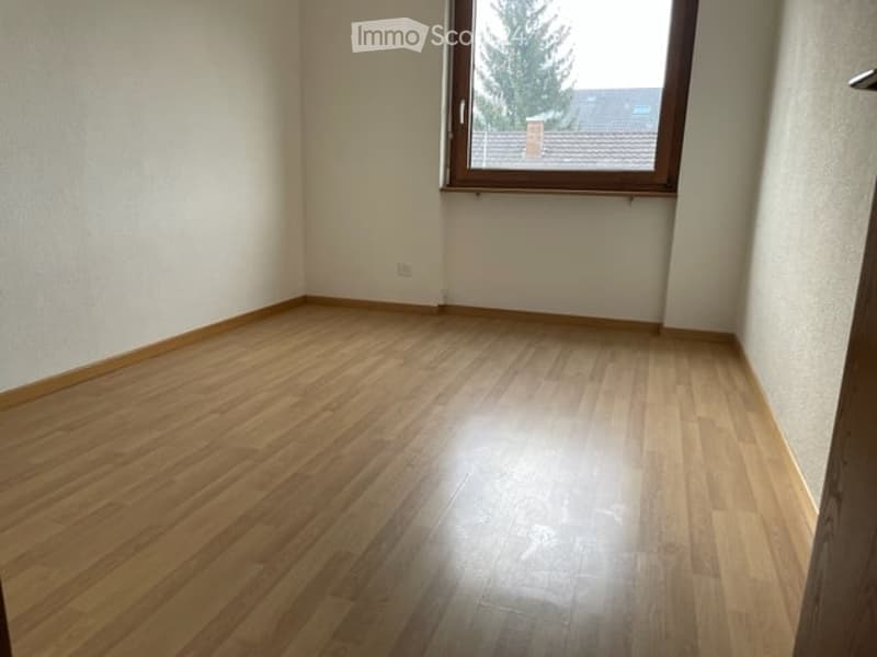 Empty room with wooden flooring and white walls, featuring a large window with a view of trees and a neighboring building.