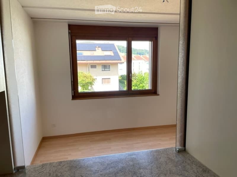 Empty room with wooden flooring, white walls, and a large window showing a view of neighboring houses and greenery.