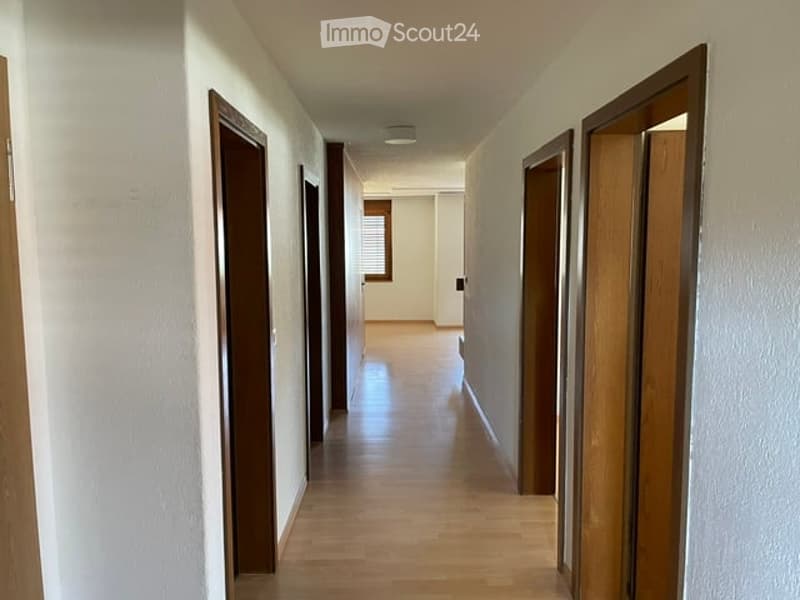 A long, narrow hallway with wooden flooring and white walls, featuring several wooden doors on both sides and a small window at the far end.