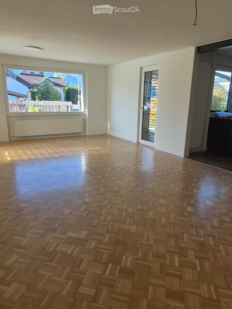 Empty room with wooden herringbone flooring, large window with view of neighboring houses, sliding glass door leading to balcony, and white walls.