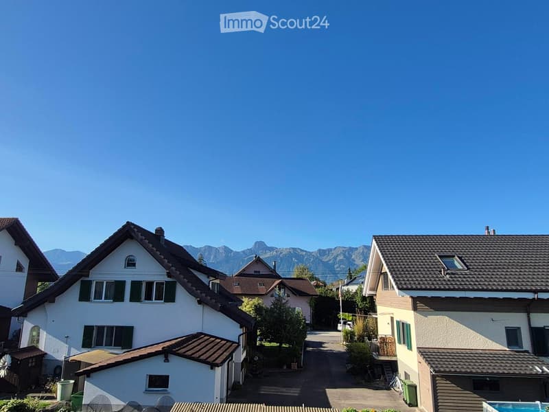 Residential street with multiple houses, surrounded by greenery and mountains in the background.
