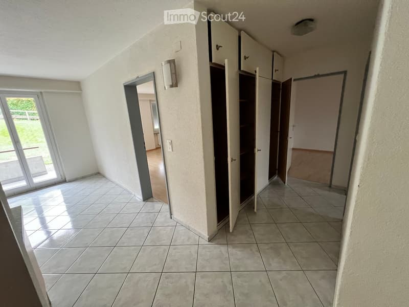 Empty hallway with tiled floor, white walls, and open doors leading to other rooms. Natural light enters through a large window on the left.
