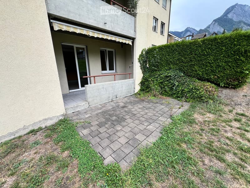 Garden area with a paved pathway leading to a building entrance, featuring a small concrete platform, a striped awning, and a view of mountains in the background.