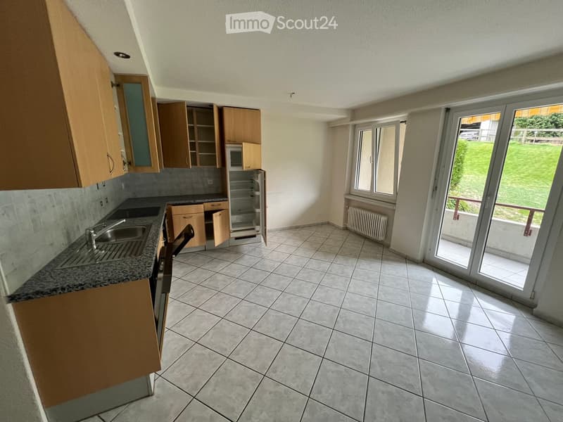 Empty kitchen with tiled floor, granite countertop, wooden cabinets, sink, microwave, and refrigerator. Large sliding glass door leads to a balcony with a view of a green lawn.