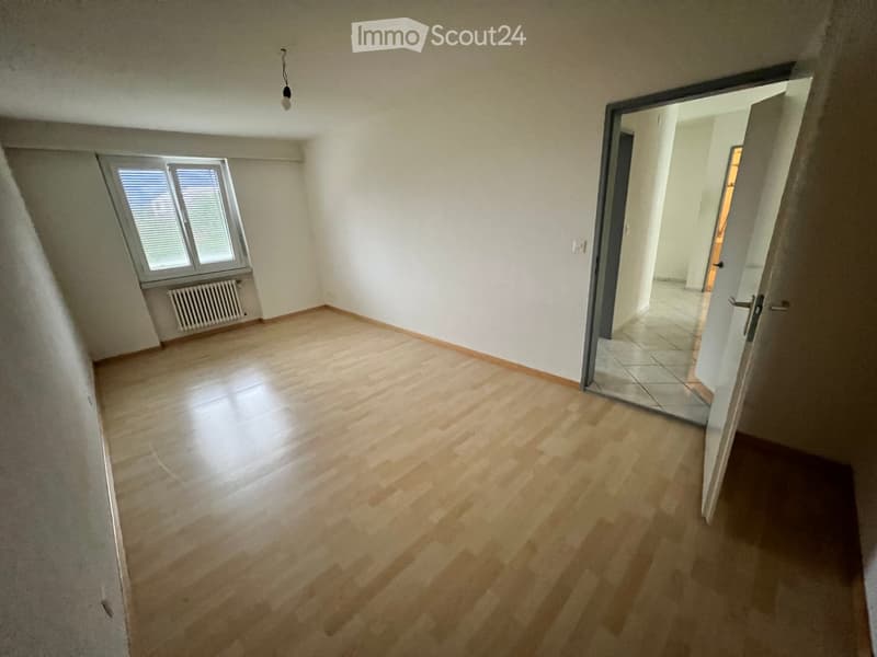 Empty room with wooden flooring, white walls, and a window with blinds. A door leads to another room with tiled flooring.