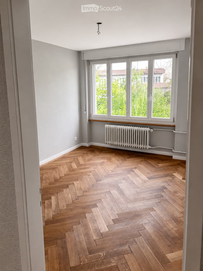 Empty room with wooden herringbone flooring, light grey walls, large windows with white frames, radiator, and visible electrical outlets.
