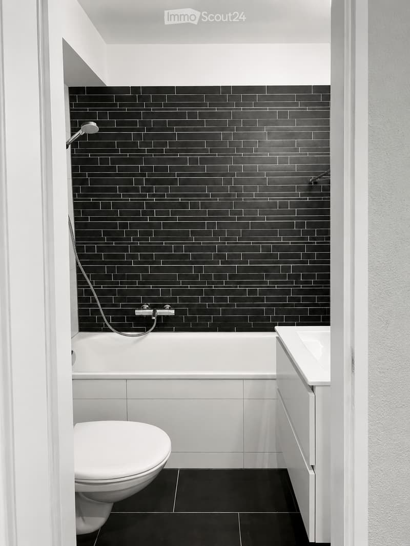 Modern bathroom with black tiled walls, white bathtub, toilet, and sink, featuring a sleek, contemporary design.