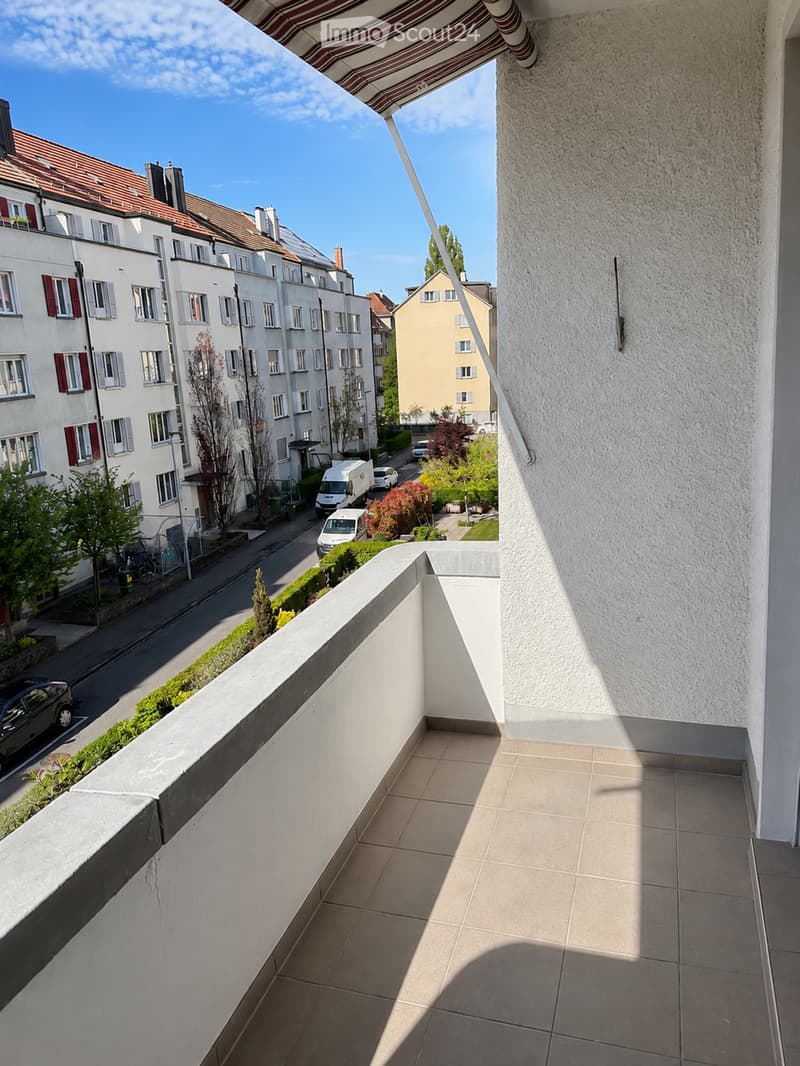 Balcony with tiled floor, white walls, and a striped awning extending over the edge. The view shows a street with buildings, parked cars, and greenery.