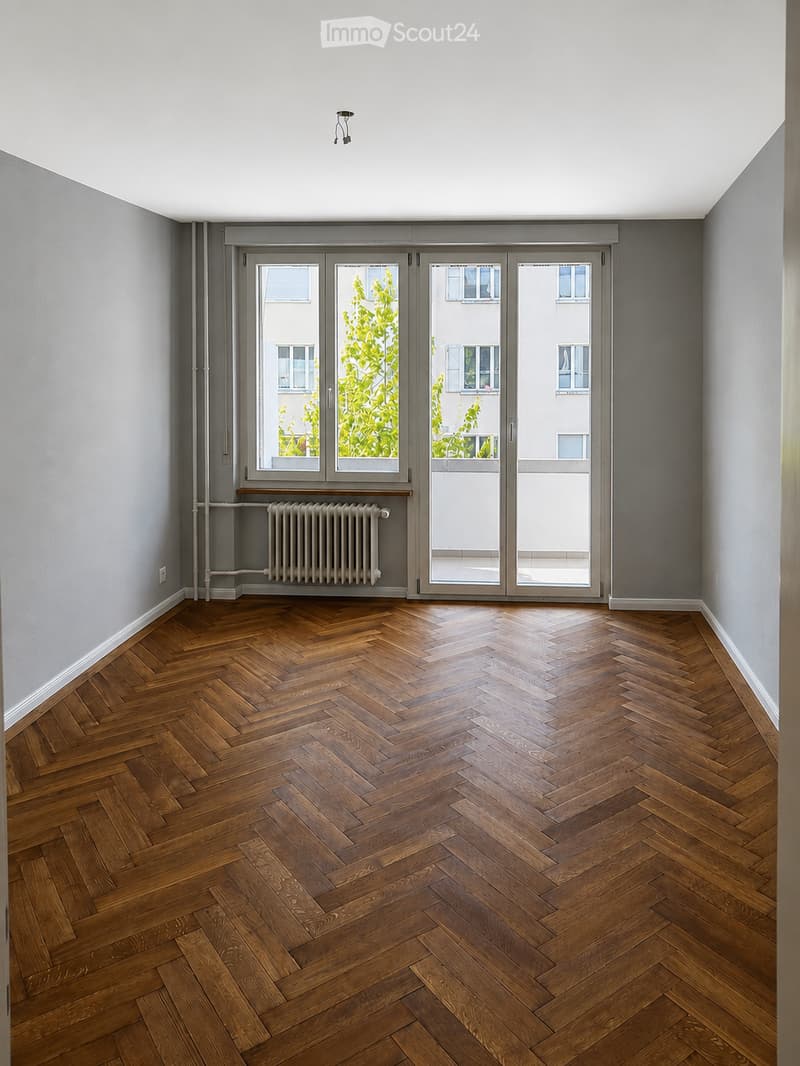 Empty room with herringbone wooden flooring, grey walls, large glass doors leading to a balcony, radiator, and visible light fixture on the ceiling.