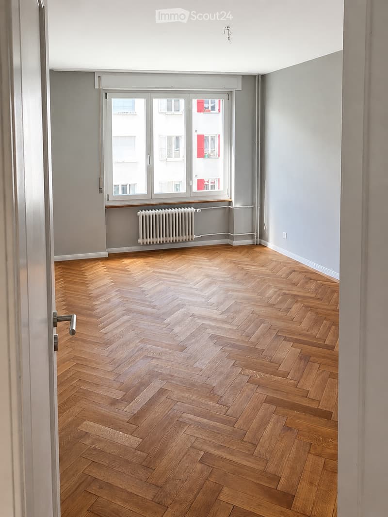 Empty room with herringbone wooden flooring, grey walls, large window with white frame, radiator, and a door with silver handle.