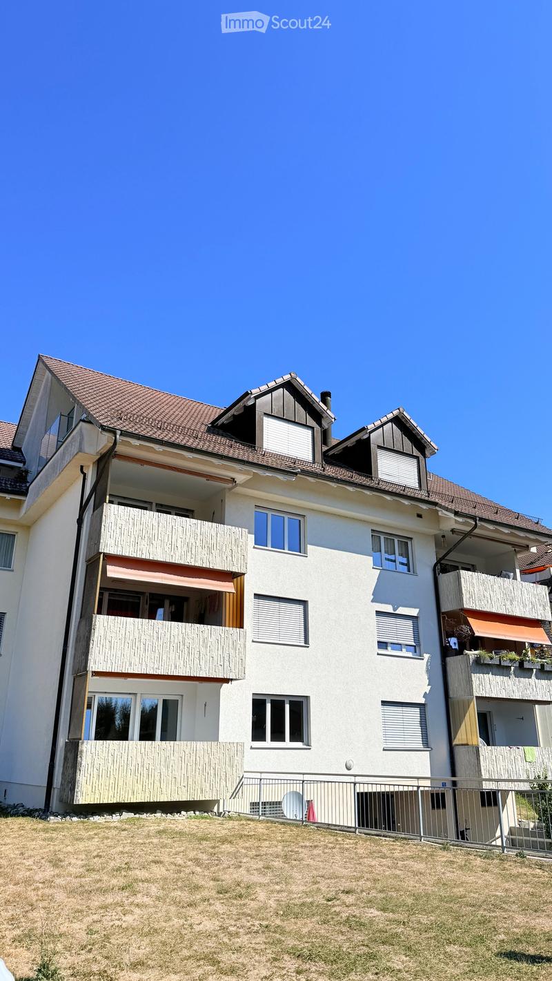 A modern, multi-story house with white walls, brown tiled roof, multiple balconies, and large windows. The house features a well-maintained lawn in front.