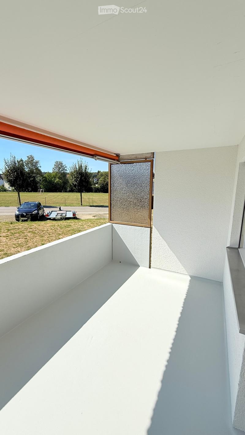 Modern white balcony with a sliding glass door, open to a grassy area with trees and a parked car.