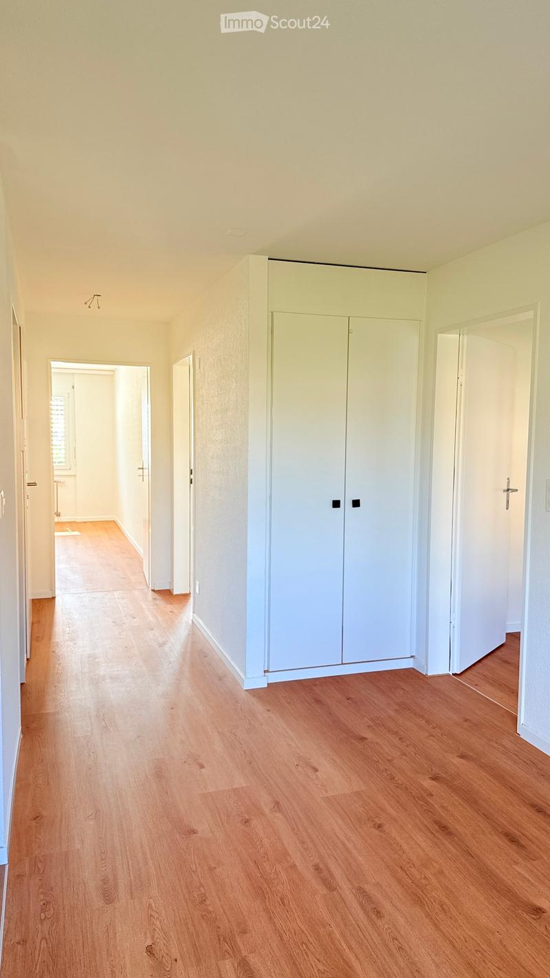 Empty hallway with light wooden flooring, white walls, and built-in white closet units on one side. The hallway leads to an open doorway revealing another room with similar flooring and light-colored walls.