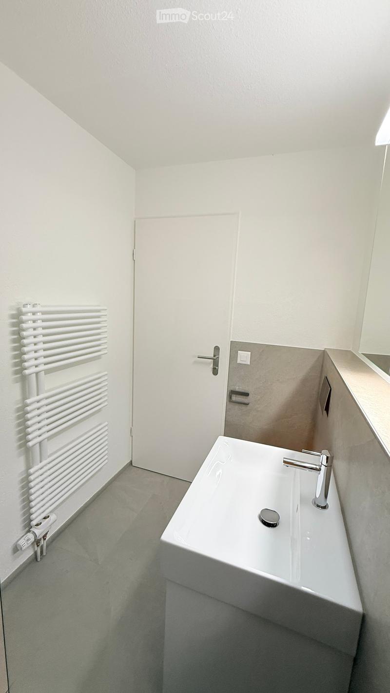 Modern bathroom with a white sink, chrome faucet, and a white radiator on the wall. The room has light-colored walls and a grey floor.