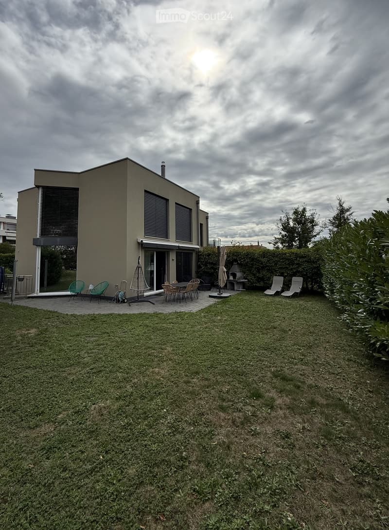 A modern house with a spacious garden area featuring a patio with chairs and a table, surrounded by greenery and a lawn.