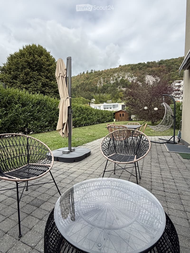 Outdoor patio with wicker chairs, glass table, umbrella, and scenic hillside view.