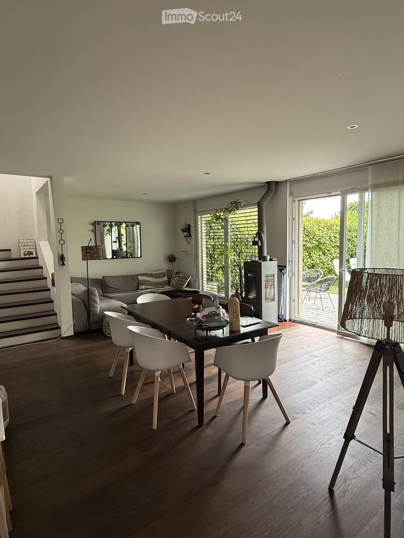 Modern living and dining area with a grey sofa, a black dining table with white chairs, a large mirror, a floor lamp, and a fireplace. The space features wooden flooring and large windows with blinds.