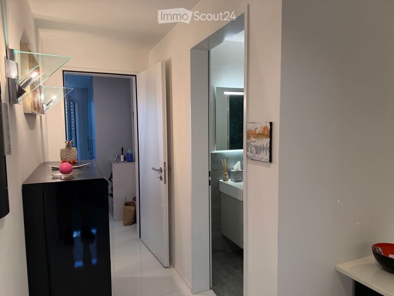 Modern hallway with white walls, tiled floor, and glass shelves on the left. An open door leads to a bathroom with a sink, mirror, and decorative items. A small table with a pink sphere and other items is visible.