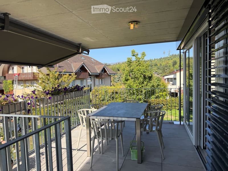 Balcony with a table and chairs, overlooking a green residential area with houses and trees.