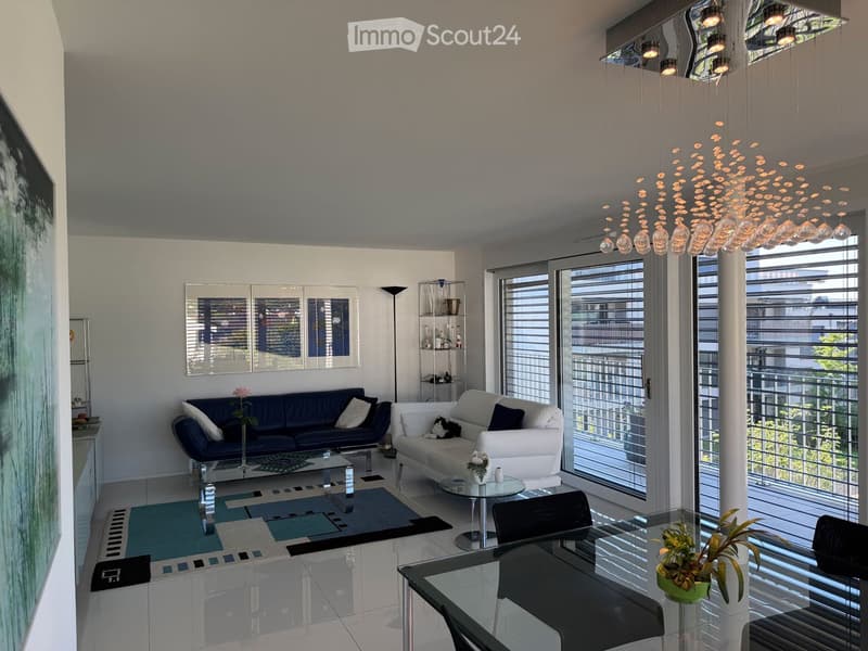 Modern living room with white walls, large windows, and a view of the city. Features a blue sofa, white armchair, glass coffee table, and a dining area with a glass table and black chairs.