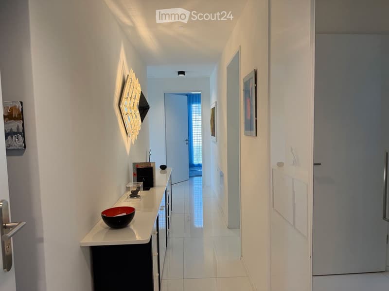 Modern hallway with white walls, sleek furniture, and artwork. Features a small console table with decorative items, a door leading to another room, and framed artwork on the walls.