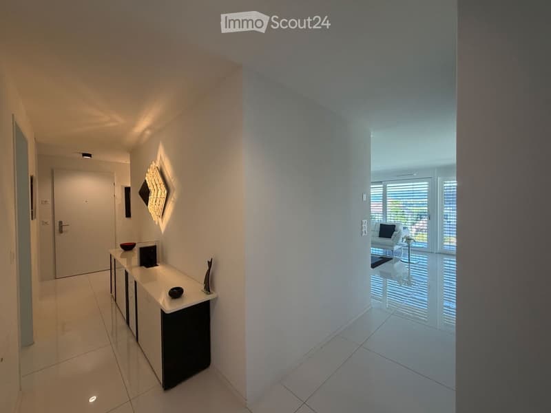 Modern hallway with white walls, tiled floor, built-in console table with decorative items, and a view into a living area with a sofa and large windows.