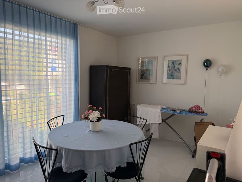 A furnished room with a round table covered in a light blue tablecloth, surrounded by black chairs. A white flower vase with pink and white flowers sits on the table. A large window with blue curtains lets in natural light. A black wardrobe is positioned