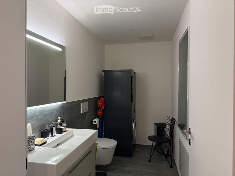 Modern bathroom with a white sink, large mirror, toilet, dark cabinet, and a small black chair near the door.