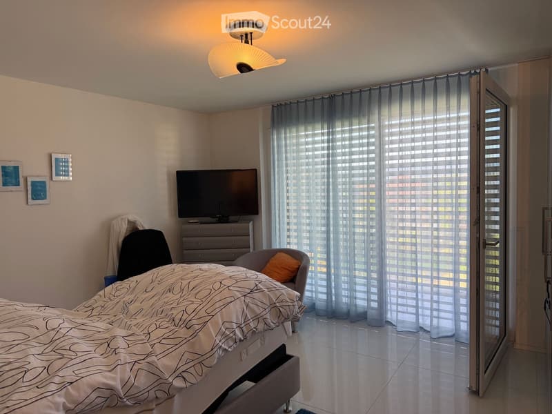 Modern bedroom with a large bed, a TV on a dresser, a chair with an orange pillow, and large windows with sheer curtains.