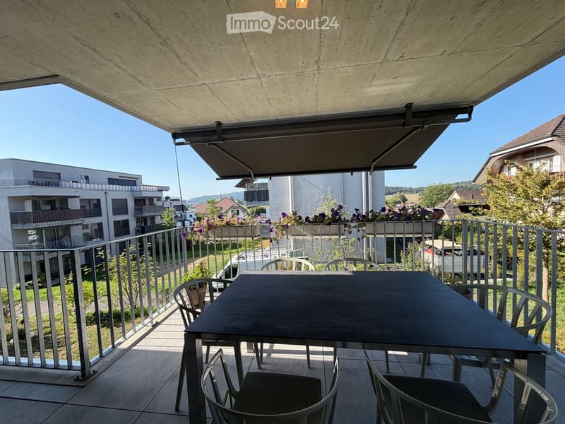 Balcony with a table and chairs, metal railing, and a retractable awning overhead. The view shows neighboring buildings and greenery.