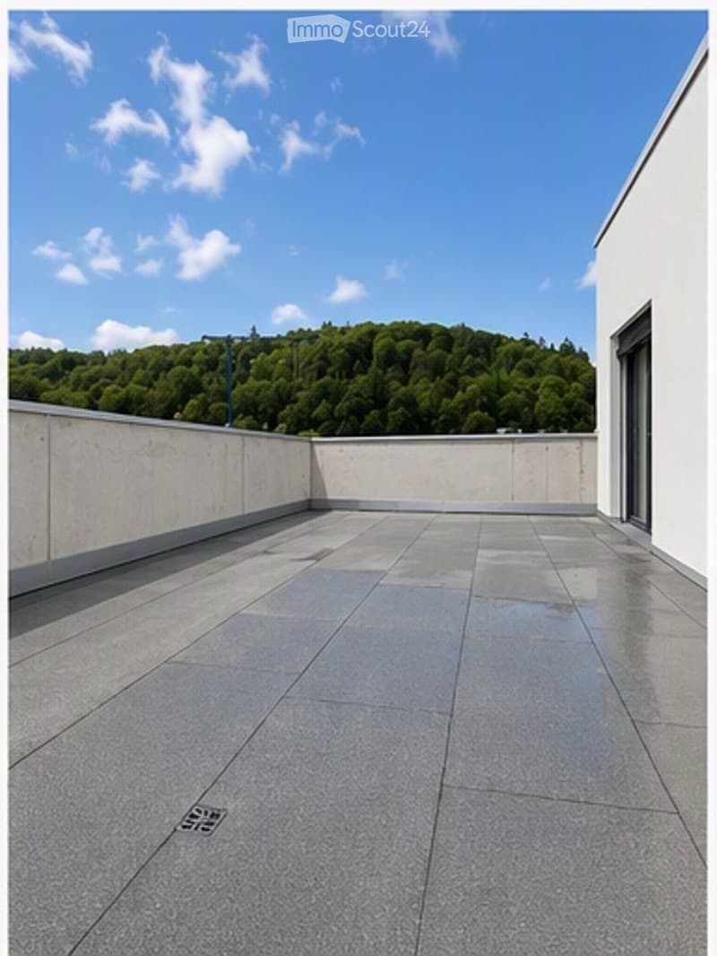 Roof terrace with smooth concrete tiles, low concrete walls, and a scenic view of a green forested hill under a partly cloudy sky.