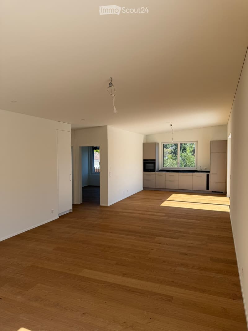 Empty room with wooden flooring, white walls, and a visible kitchen area with cabinets and appliances in the background.