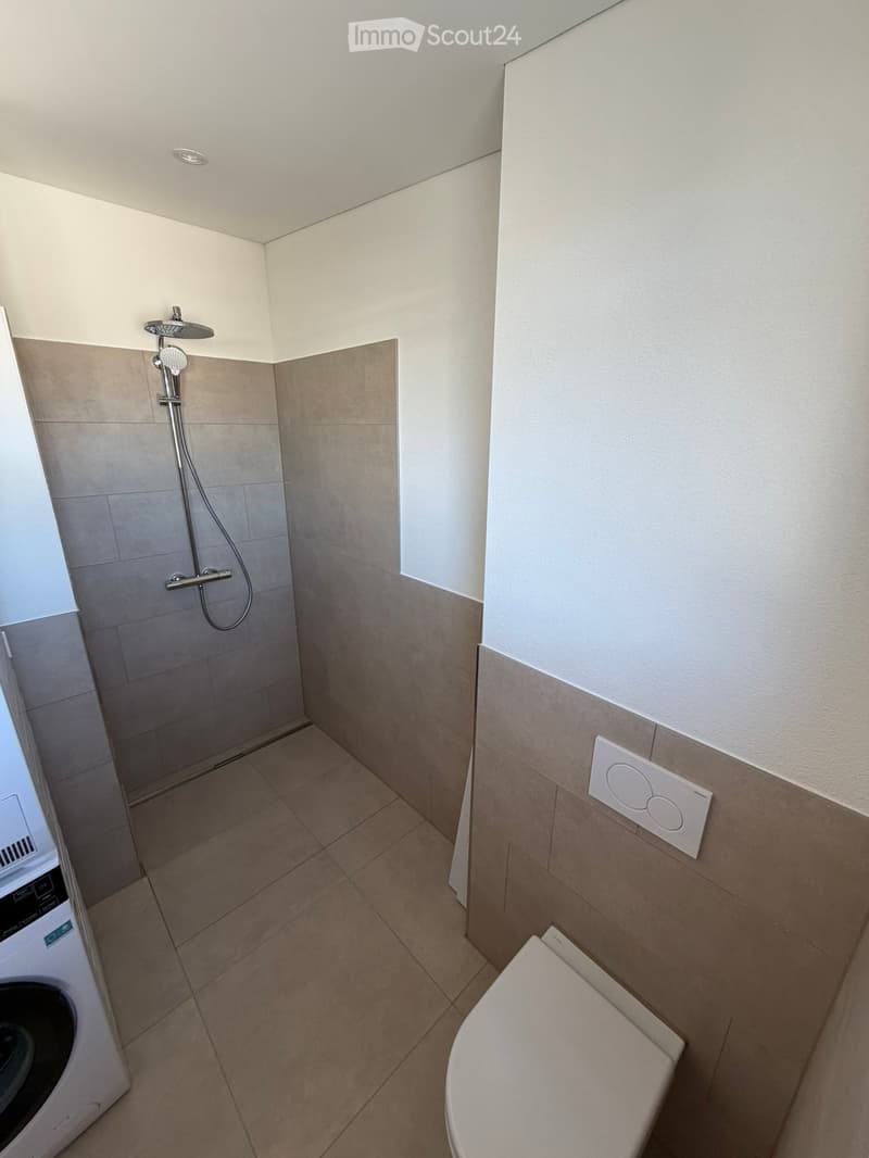 Modern bathroom with a toilet, shower, and washing machine. Light-colored tiles and walls, with a sleek showerhead and control panel.
