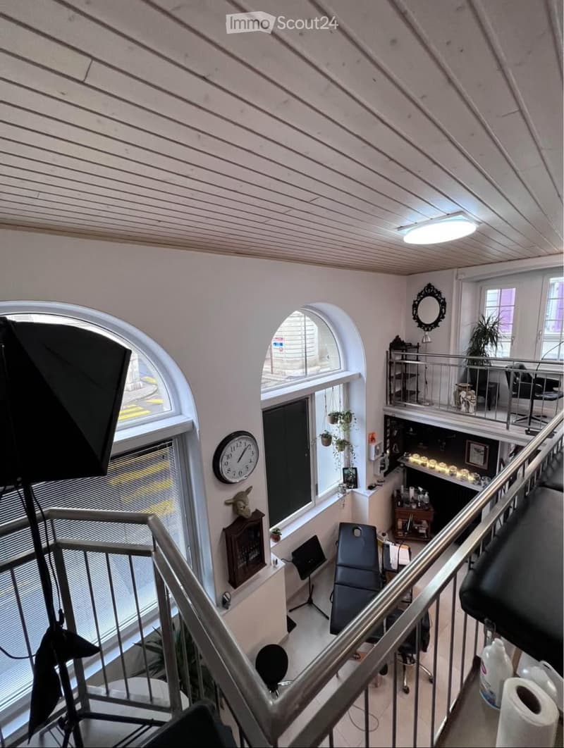 A multi-level interior space with a staircase, large windows, a chair, a small table, a mirror, a clock, and plants.