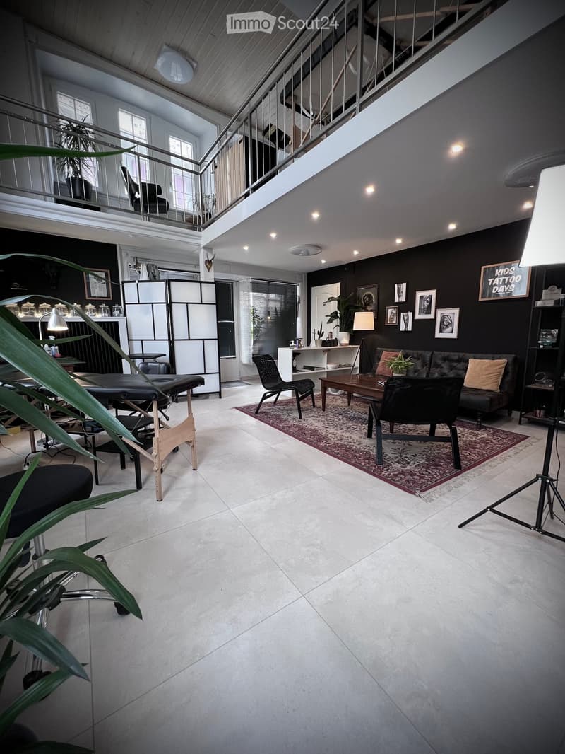 Modern living room with dark walls, white tiled floor, large windows, and a balcony. Features a black leather sofa, coffee table, chairs, a small table with a lamp, and decorative plants.