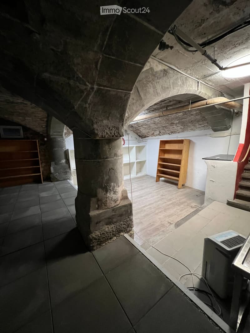 A room with stone walls and a wooden floor, featuring a large stone pillar, a wooden shelf, a small table, and a screen on a stand.