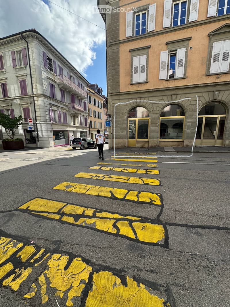 Multi-story urban buildings with classic European architecture, including balconies and shutters, lining a street with painted yellow markings on the pavement.
