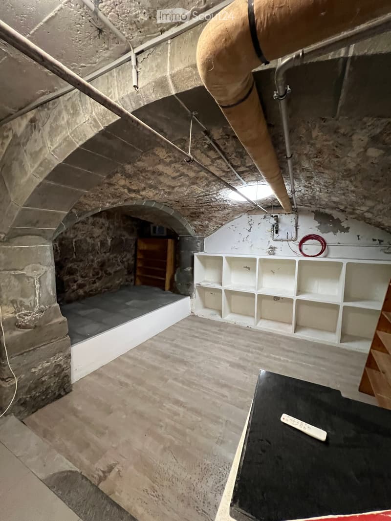 A partially furnished room with exposed stone walls and a raised platform, featuring a white shelving unit, a small table with a remote control, and a wooden shelf in the background.