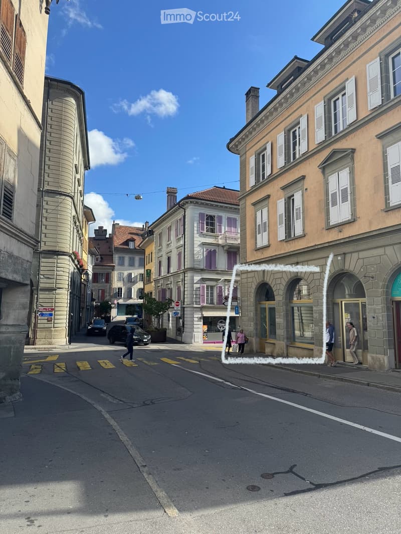 A street view of a European town with multi-story buildings, classic architecture, and a clear blue sky. The buildings feature ornate facades, shutters, and balconies.